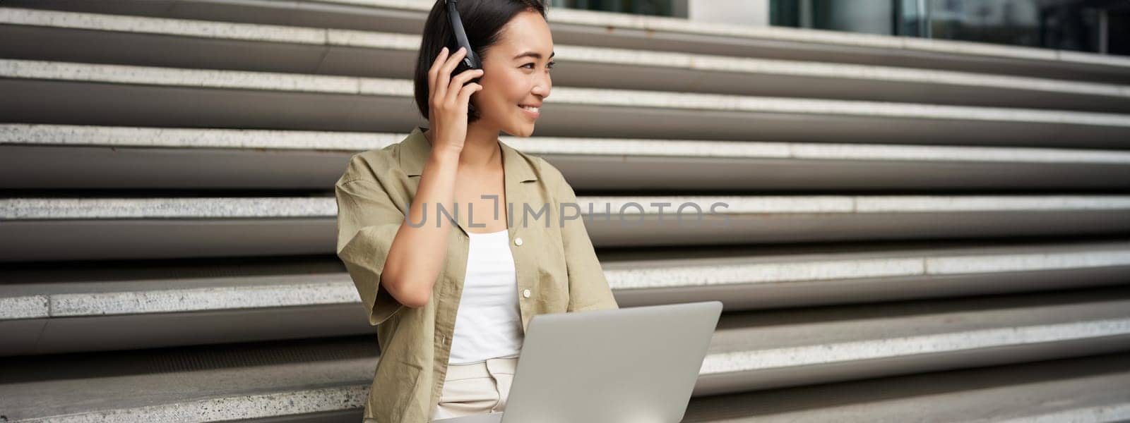 Portrait of stylish young woman smiles, sits on stairs with headphones and laptop, works on homework for university.
