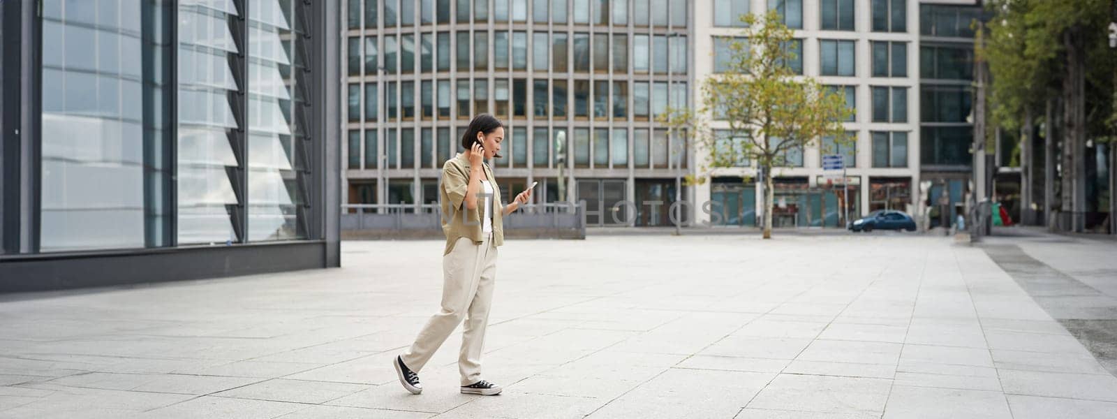 Silhouette of asian woman walking on street in wireless headphones, holding smartphone.