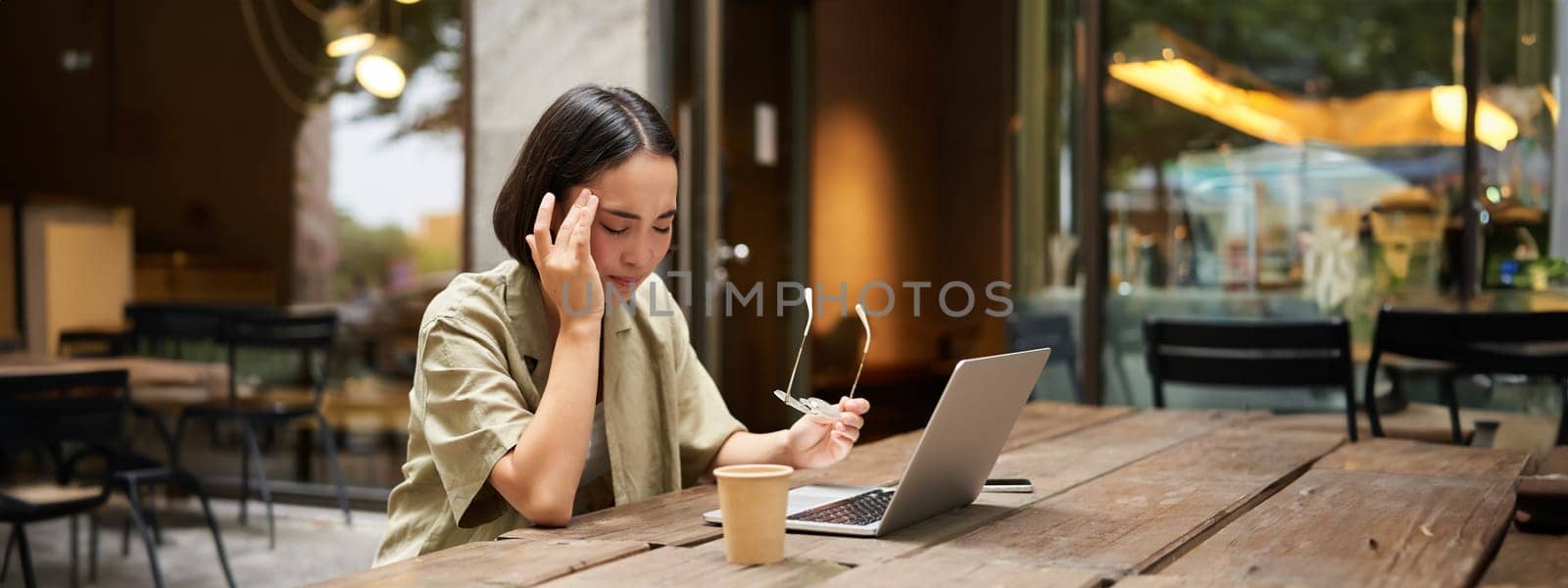 Young asian woman feeling tired after working with laptop, sitting in cafe on bench outdoors, drinking coffee, looking exhausted.