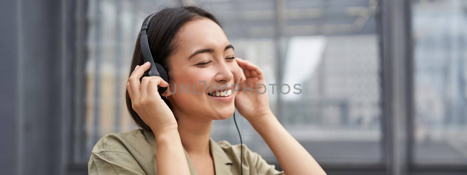 Portrait of beautiful asian woman in headphones, listening music on street of city centre, smiling happily.