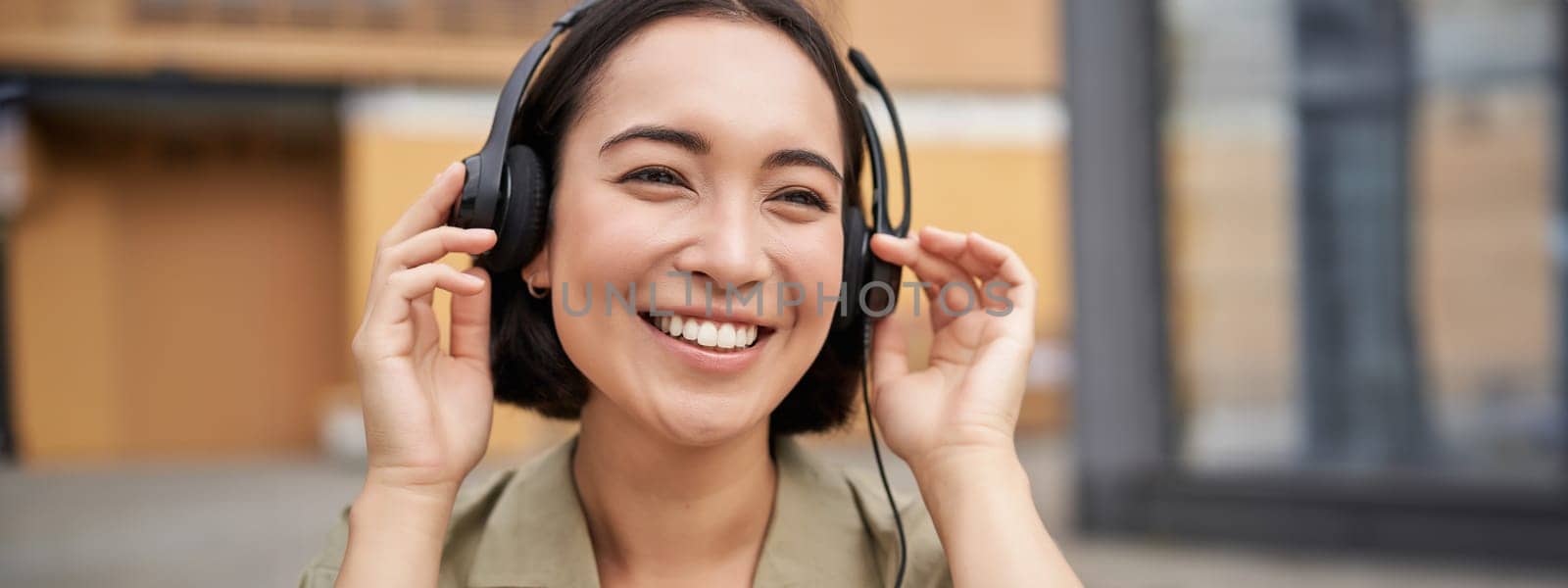 Portrait of beautiful asian woman in headphones, listening music on street of city centre, smiling happily.