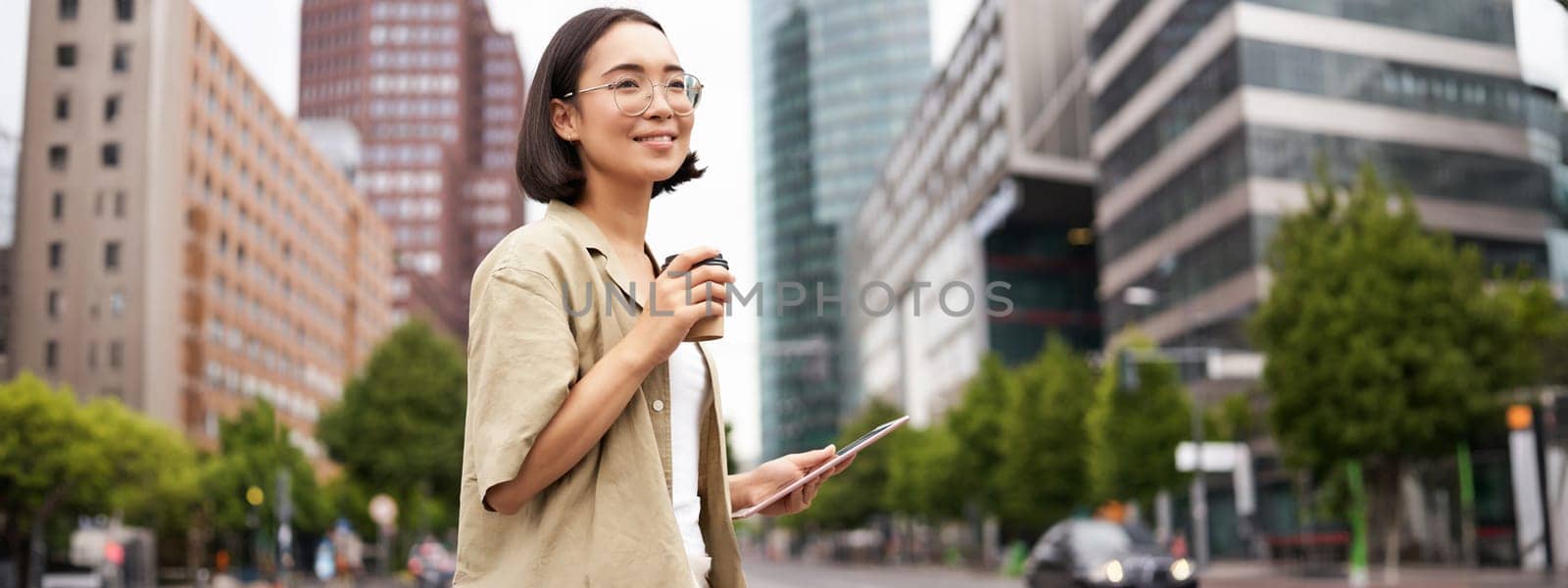 Happy woman exploring the city. Young korean girl holds tablet, drinks coffee and walks along street with big smile on her face.