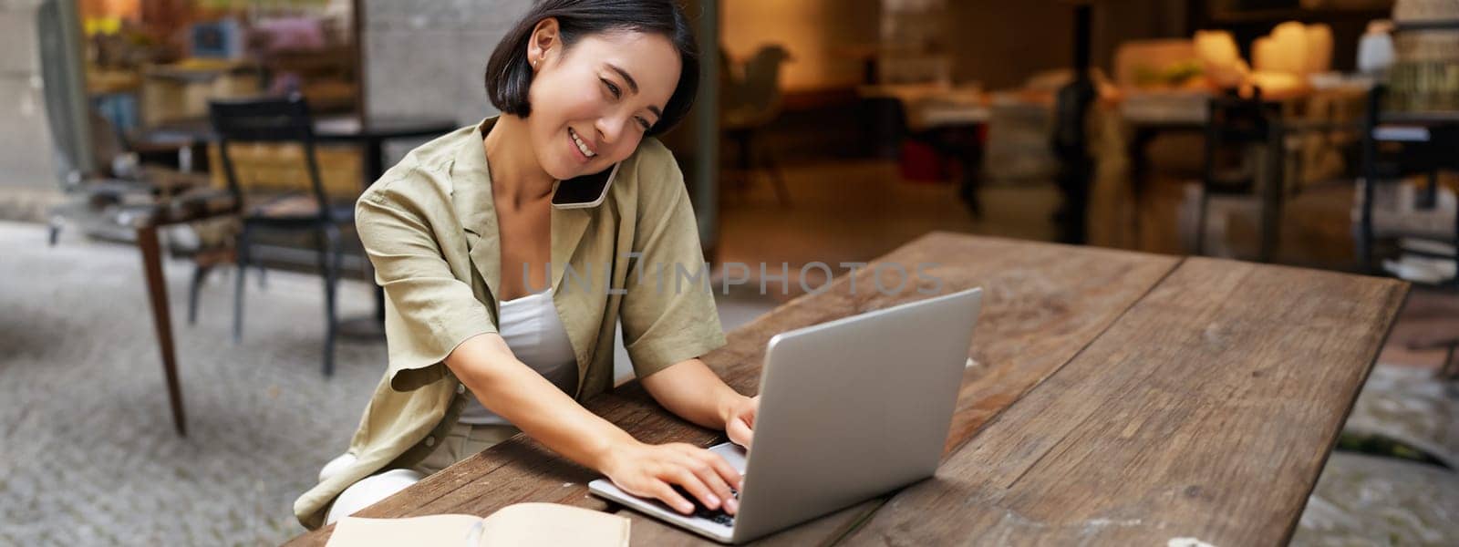 Portrait of young woman talking on mobile phone while typing, using laptop, working remotely from outdoo cafe, doing homework and calling.