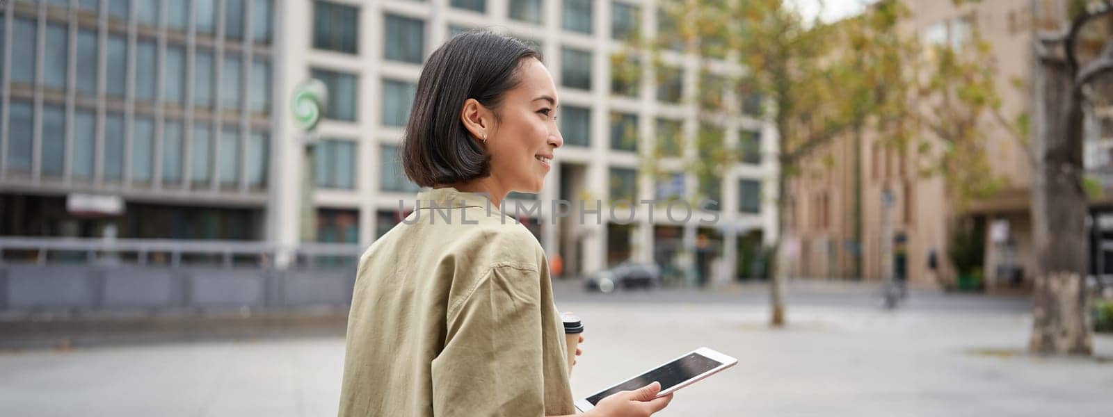 Portrait of stylish urban girl walks in city with tablet, drinks takeaway coffee.