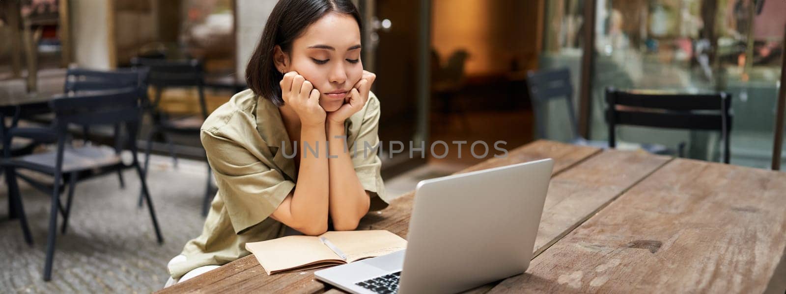 Bored girl looks at her laptop, sits outdoors in cafe, listening boring online meeting, working and feeling sad.