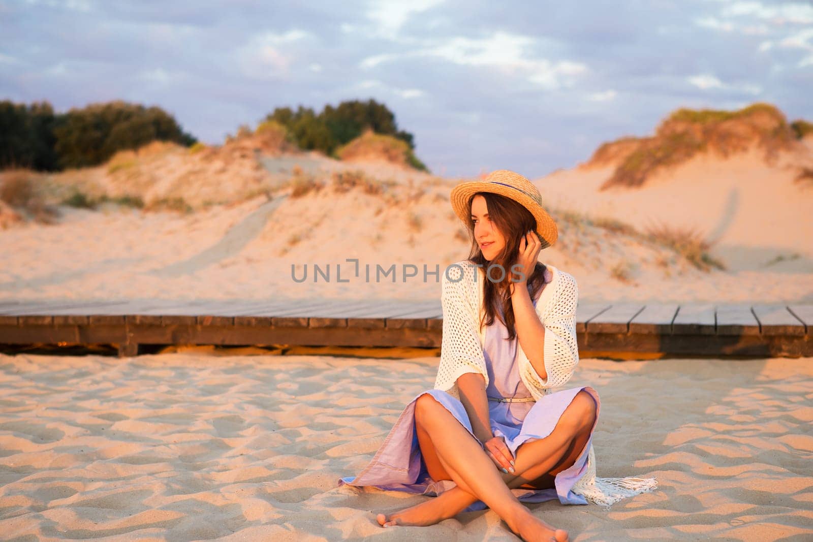 happy beautiful girl in a dress sits on the sand and looks at the sunrise.