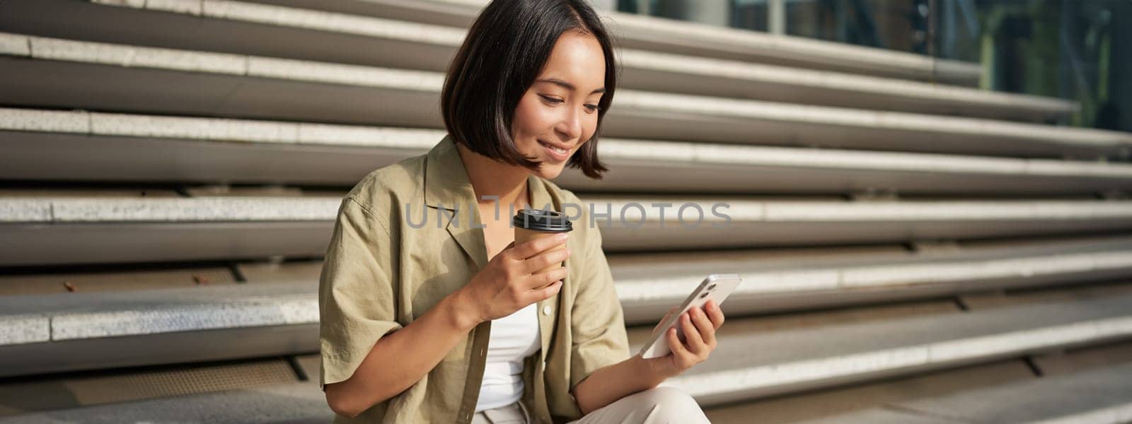 Portrait of asian woman with smartphone, drinks coffee and watches videos on mobile phone. Girl with telephone sits on stairs outdoors.