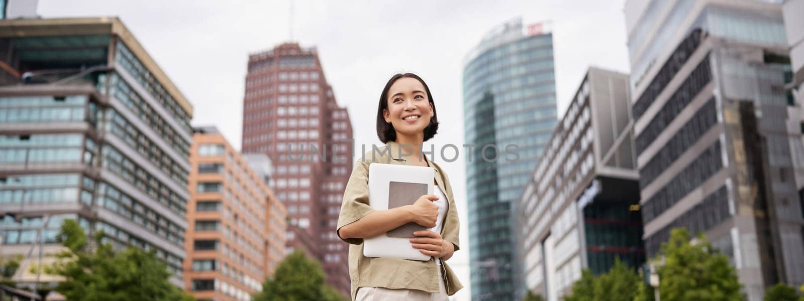 Portrait of young asian woman, looking happy and confident, going to work or university, city skyscrappers behind her, holding laptop and notebook by Benzoix