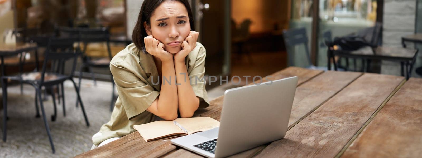 Bored girl looks at her laptop, sits outdoors in cafe, listening boring online meeting, working and feeling sad.
