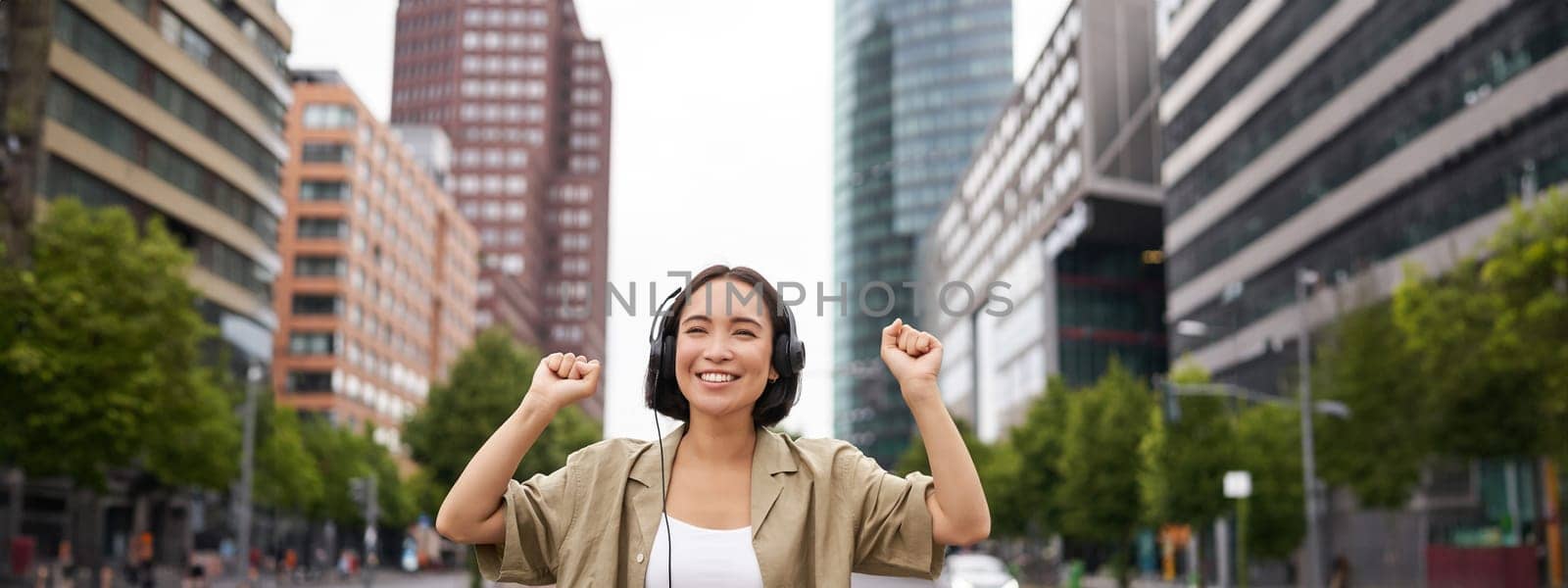 Portrait of smiling asian woman dancing, triumphing, feeling happy while listening music in city, posing on street near skyscrappers.