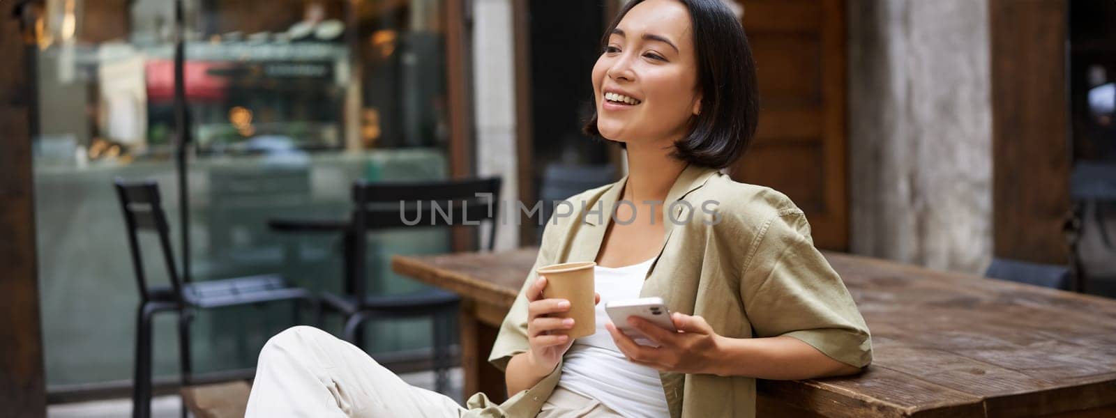Stylish urban girl sits with her phone in cafe, drinks coffee and chatting, browsing social media on smartphone.