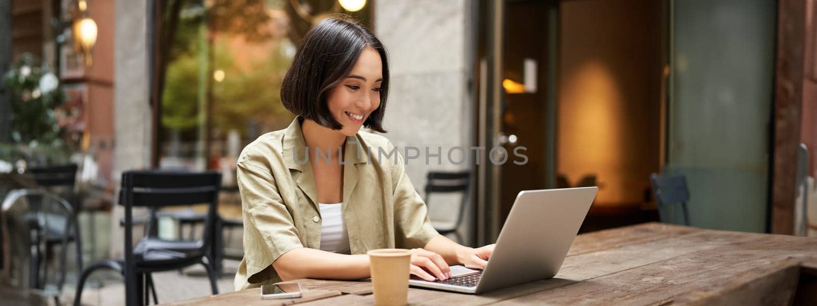 Young asian woman, digital nomad working remotely from a cafe, drinking coffee and using laptop, smiling by Benzoix