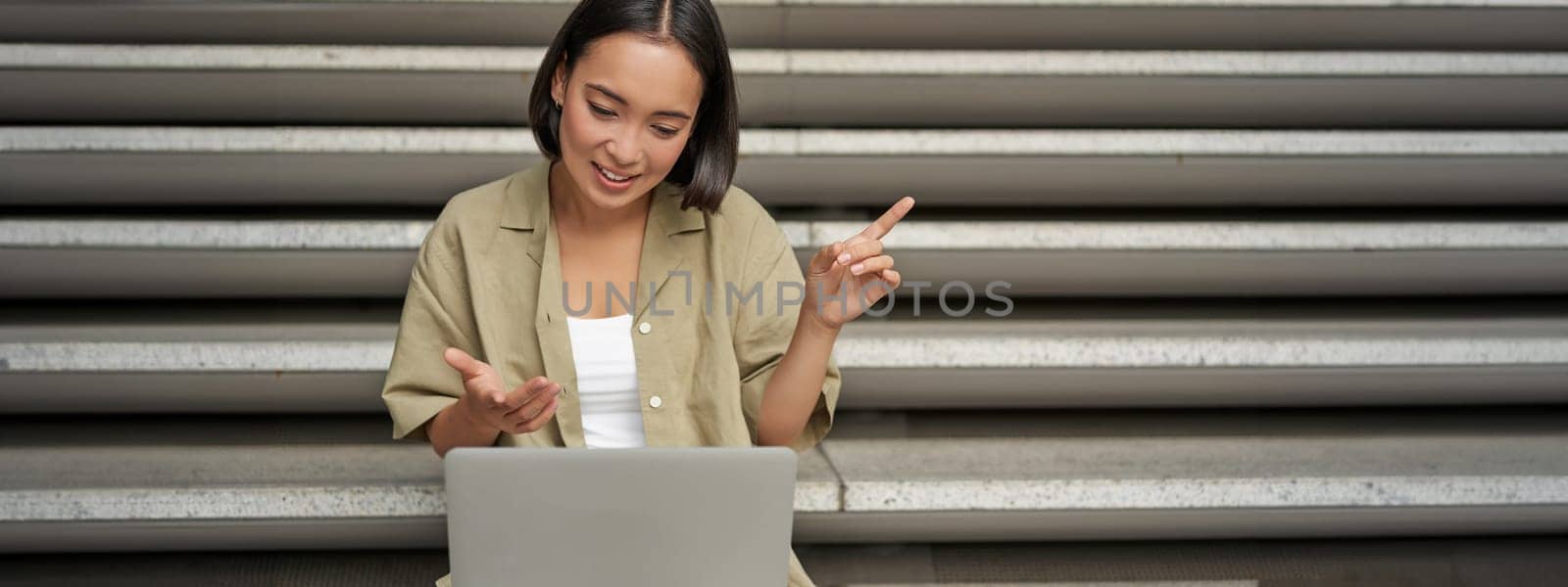 Portrait of young asian girl, student talks at laptop, video chat, speaking during online meeting, sitting outdoors on stairs by Benzoix