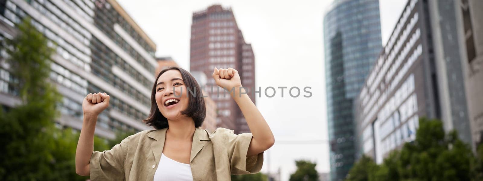 Happy people. Young smiling asian woman dancing in city, raising hands up in city, triumphing, celebrating victory.