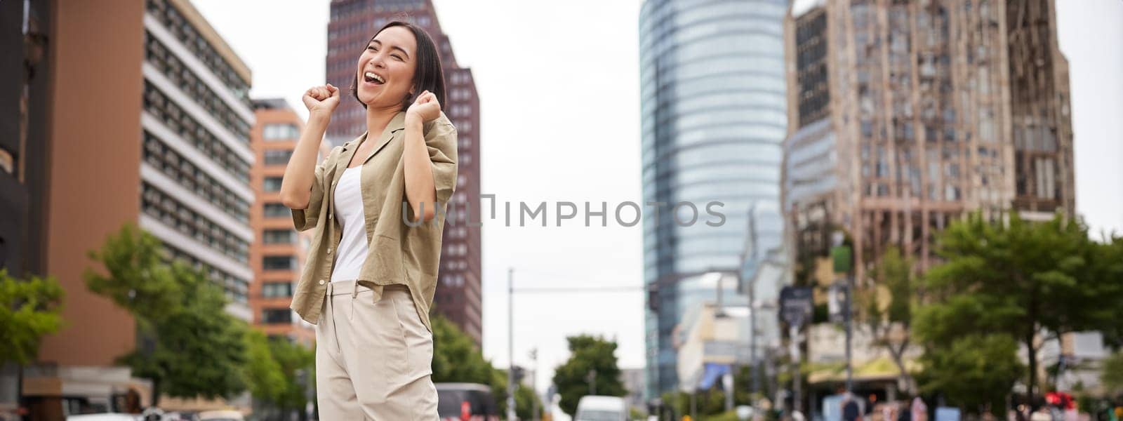 Vertical shot of young asian woman posing happy, raising hands up and dancing, triumphing, celebrating victory, enjoying day out in city.