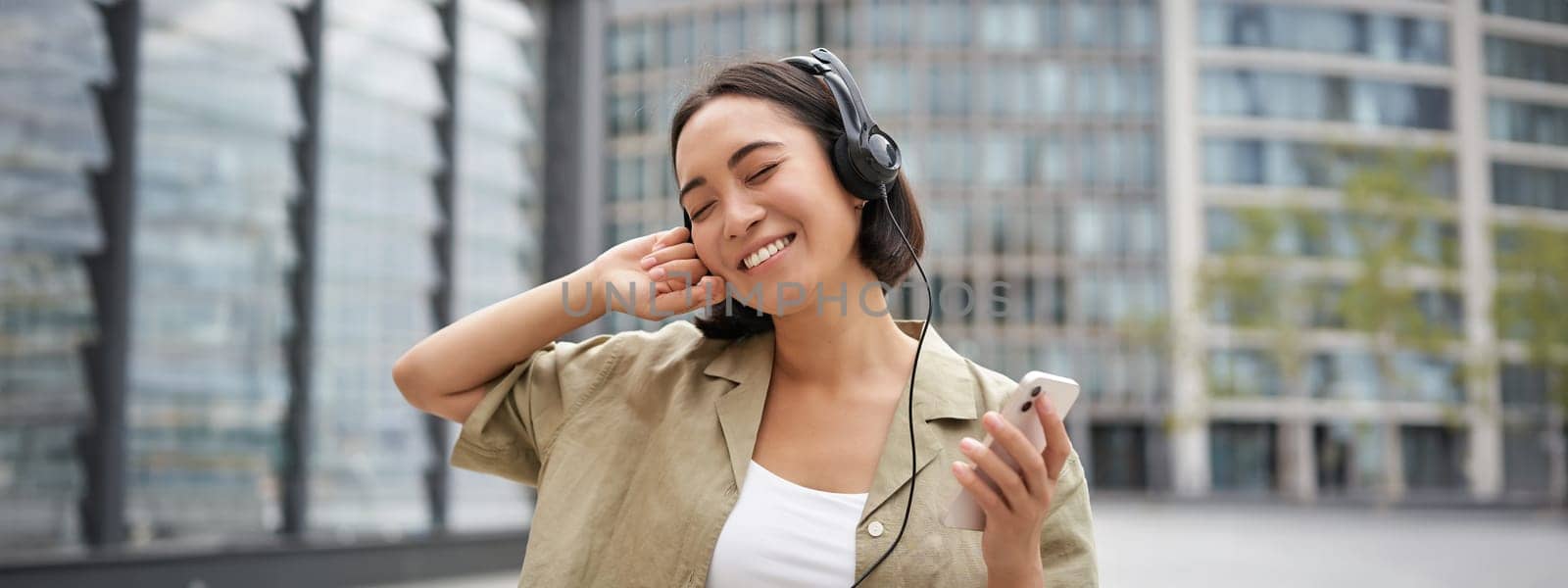 Happy young woman dancing on streets and listening music in headphones, holding smartphone.
