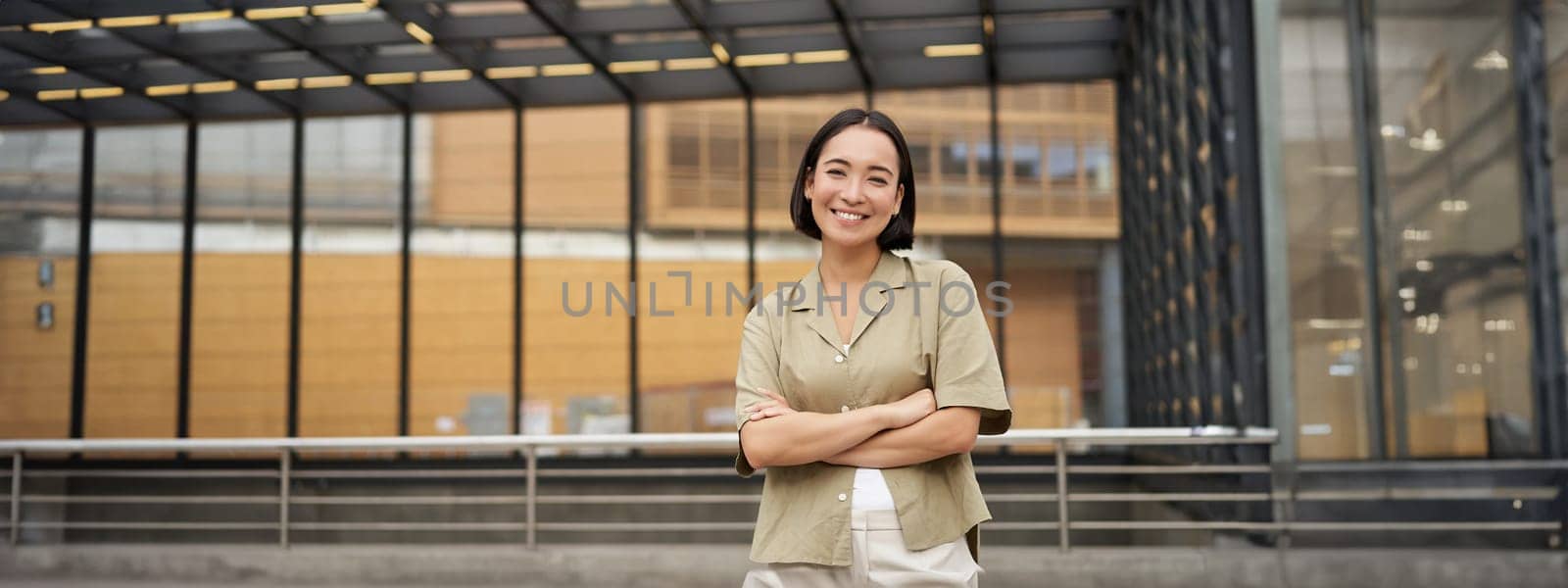 Portrait of young asian woman standing with confidence, cross arms on chest and smiling, posing outdoors on street.