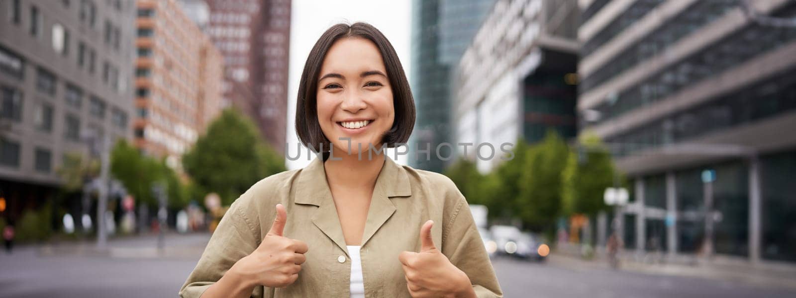 Enthusiastic asian woman , shows thumbs up in approval, looking upbeat, say yes, approves and agrees, stands on street.