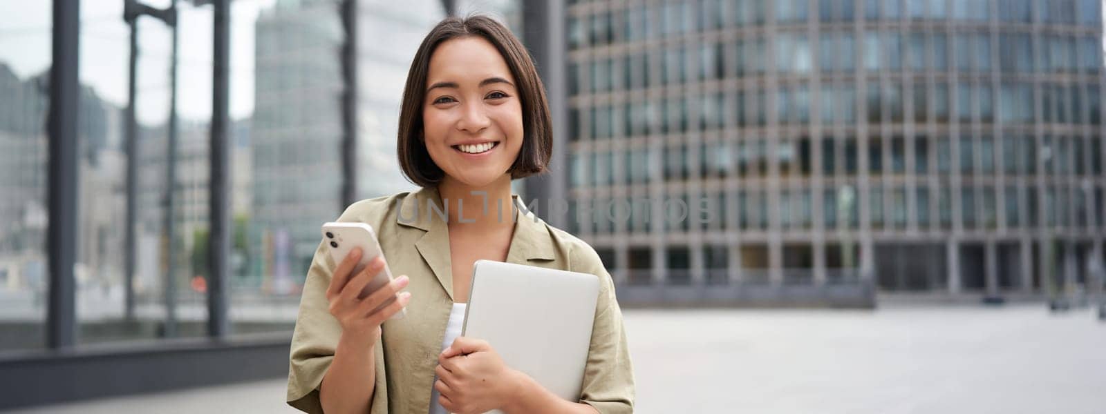 Portrait of smiling asian girl with laptop, holds mobile phone and looks happy at camera, stands on street.