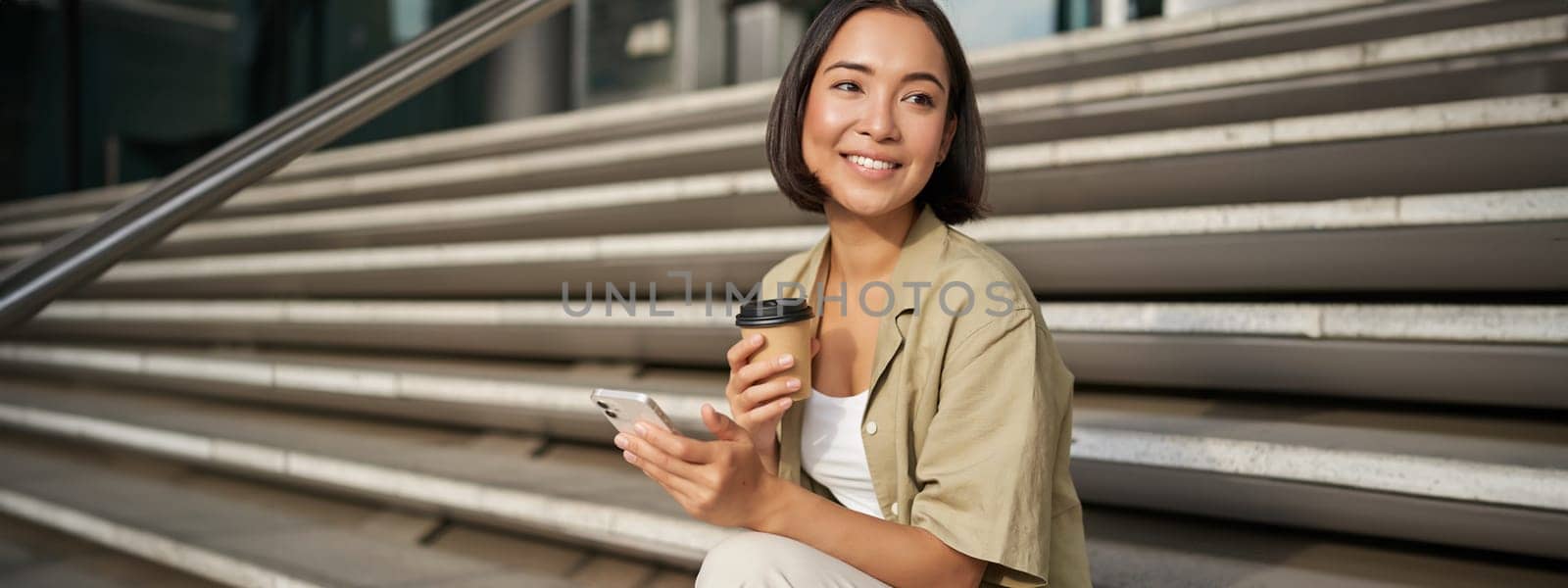 Happy urban girl drinks her takeaway coffee and scrolls feed on smartphone. Asian woman sits on stairs with tea and holding mobile phone.