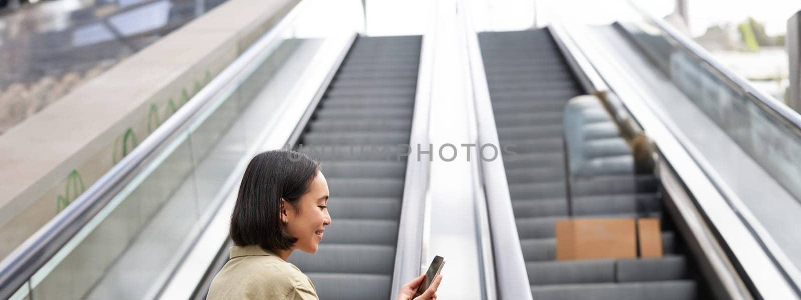 Young asian girl going up on an escalator, holding smartphone, smiling while walking in city. Copy space
