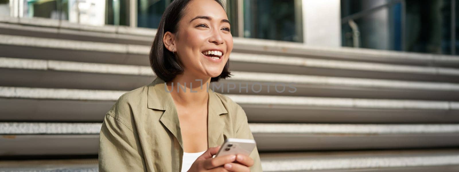 Beautiful asian girl sits on stairs with smartphone. Young korean woman resting outdoors, using mobile phone application by Benzoix