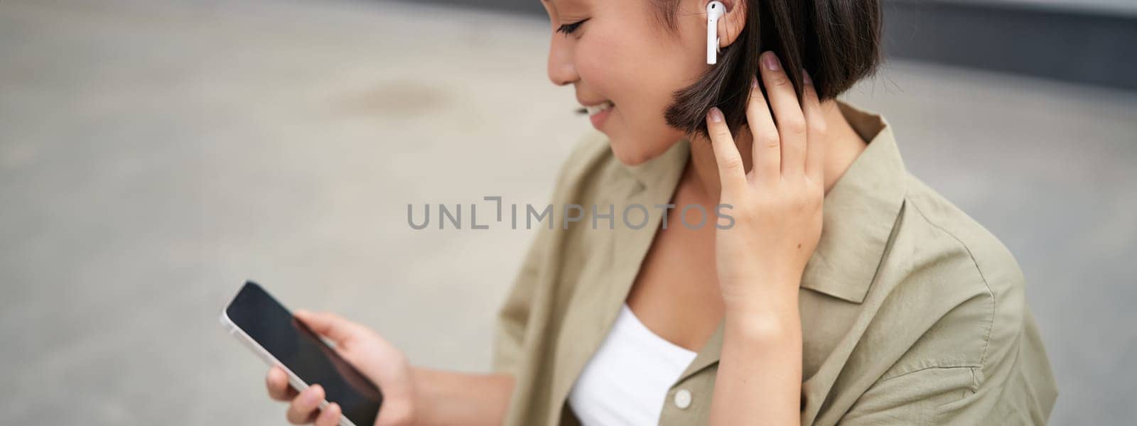 Close up portrait of asian girl, looking at mobile screen, listening music in headphones. Woman with earphones walks on street.