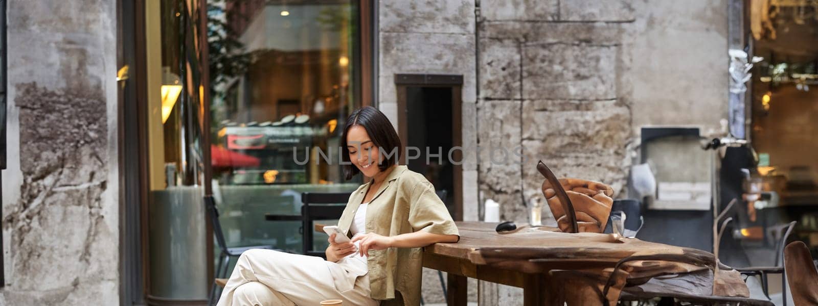 Portrait of stylish young korean woman sits in cafe, holds smartphone, smiles, enjoys coffee outdoors by Benzoix