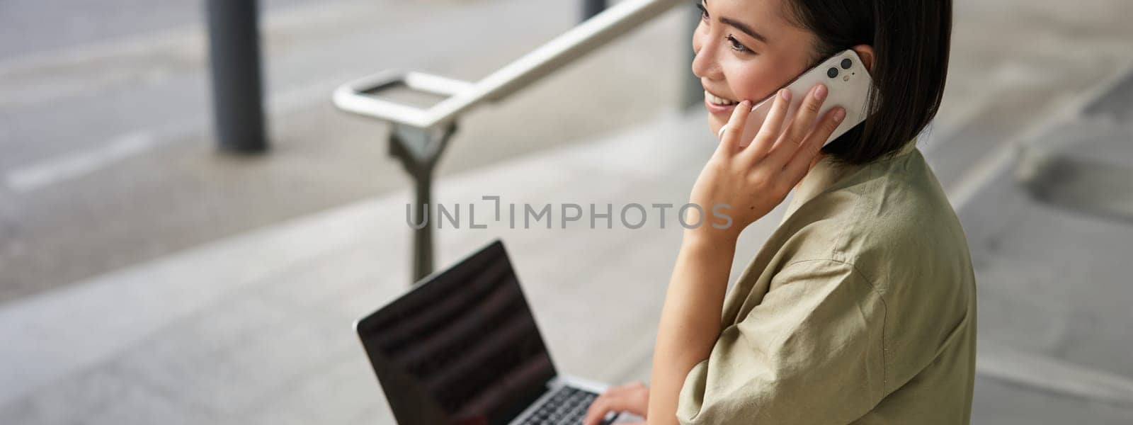 Portrait of asian girl, student sits on stairs with laptop, talks on mobile phone. Young woman makes a telephone call while working on computer.