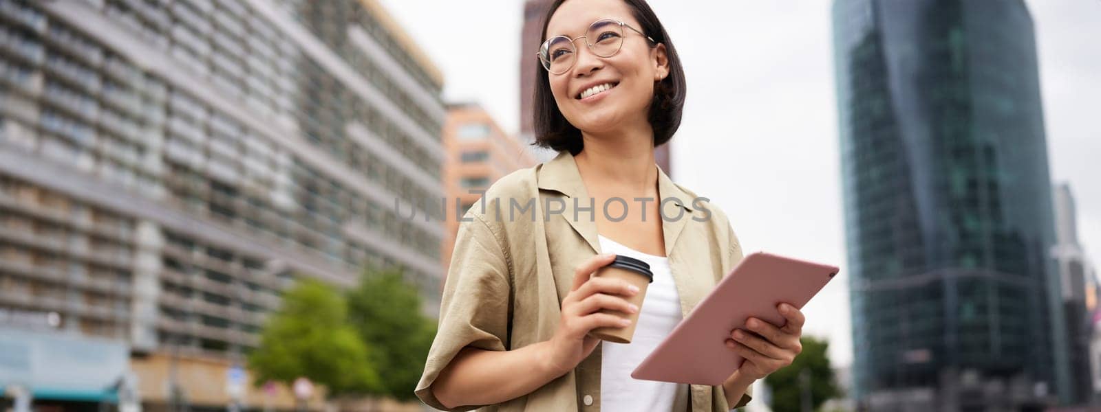 Portrait of happy young woman in glasses, standing on street with cup of coffee and tablet.