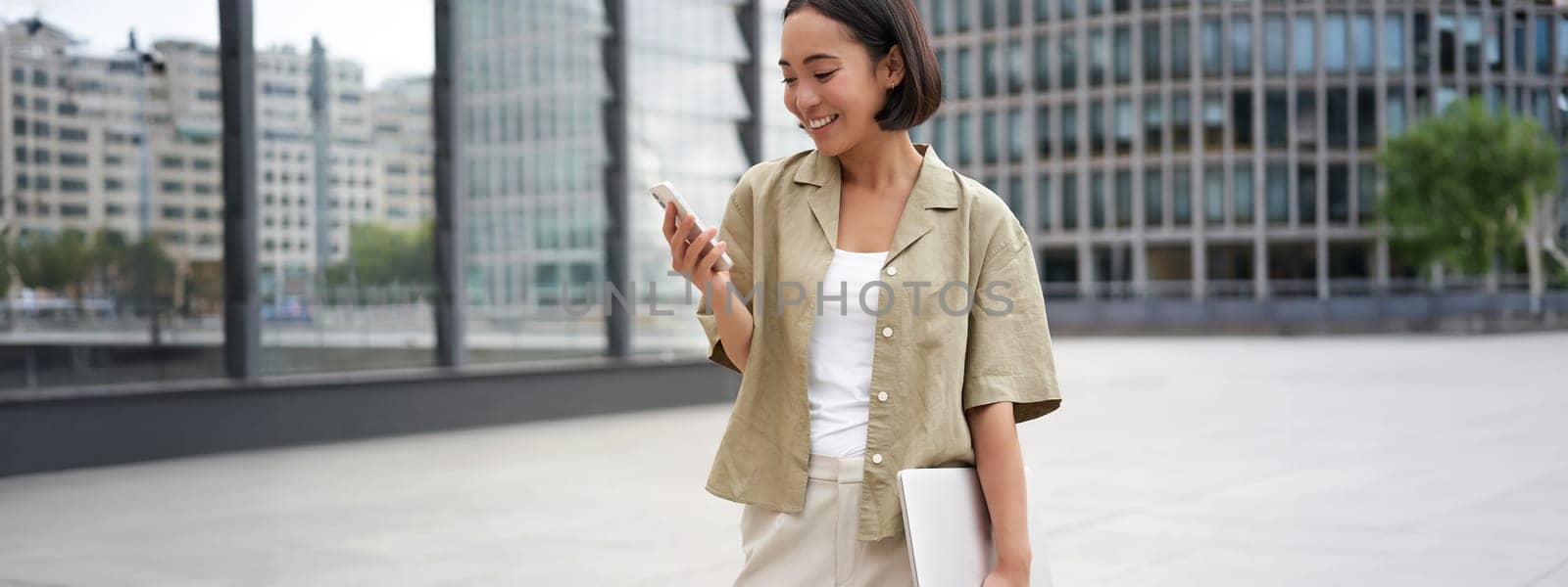 Young asian woman with laptop, walking on street and texting message, smiling while looking at smartphone by Benzoix