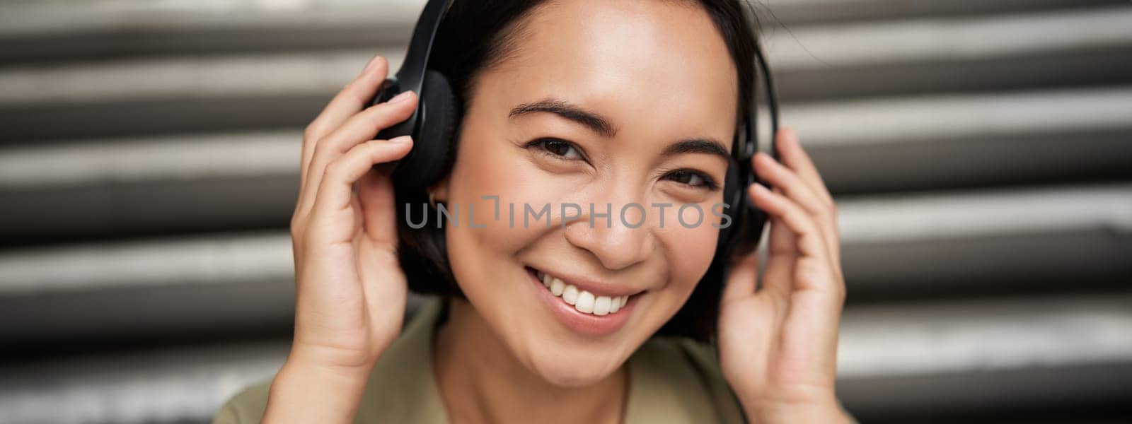Close up portrait of smiling asian girl in headphones, listens to music outdoors, looking happy. by Benzoix