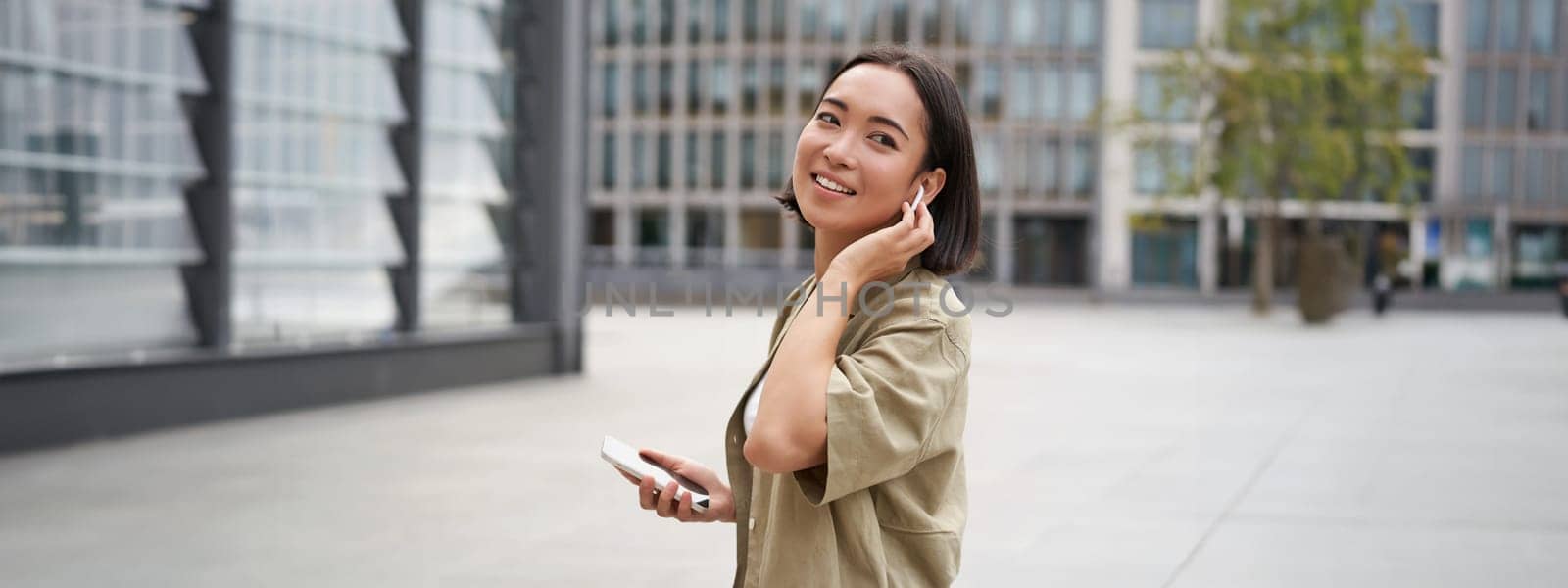 Modern people. Young woman walks on street of city and listens music in wireless headphones, holds smartphone.