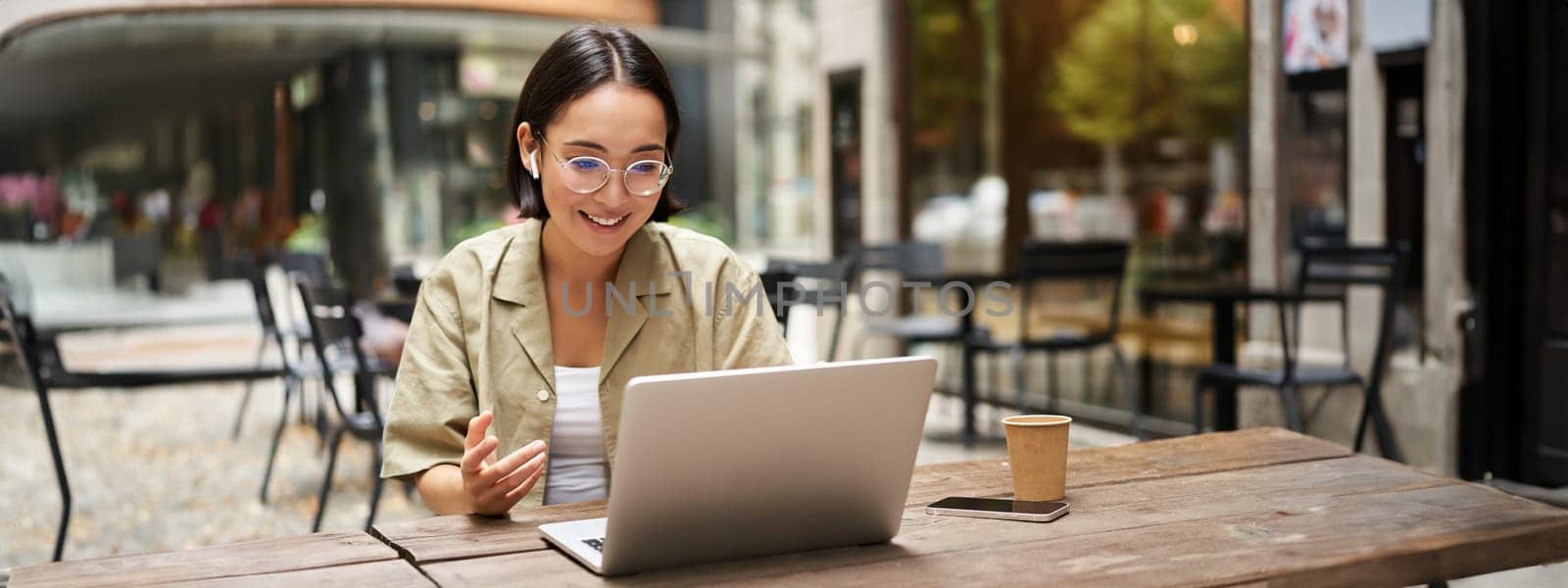 Young woman sitting on online meeting in outdoor cafe, talking to laptop camera, explaining something, drinking coffee by Benzoix