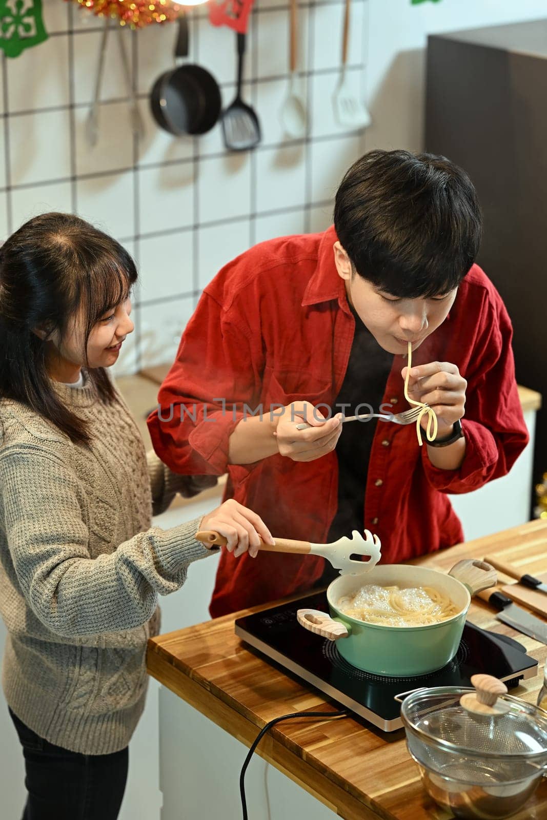 Young couple tasting freshly cooked pasta in a warm Christmas kitchen.