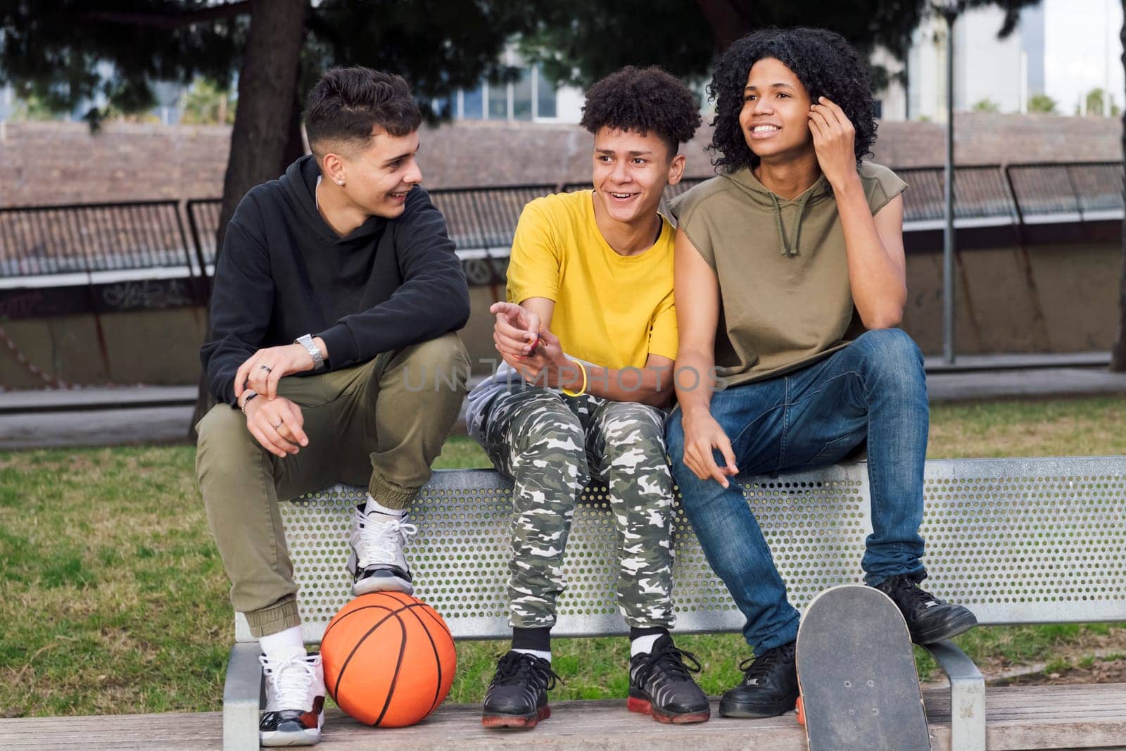 multiracial teen male friends smiling happy while talking sitting on bench of a park with basketball and skateboard, concept of youth and urban lifestyle