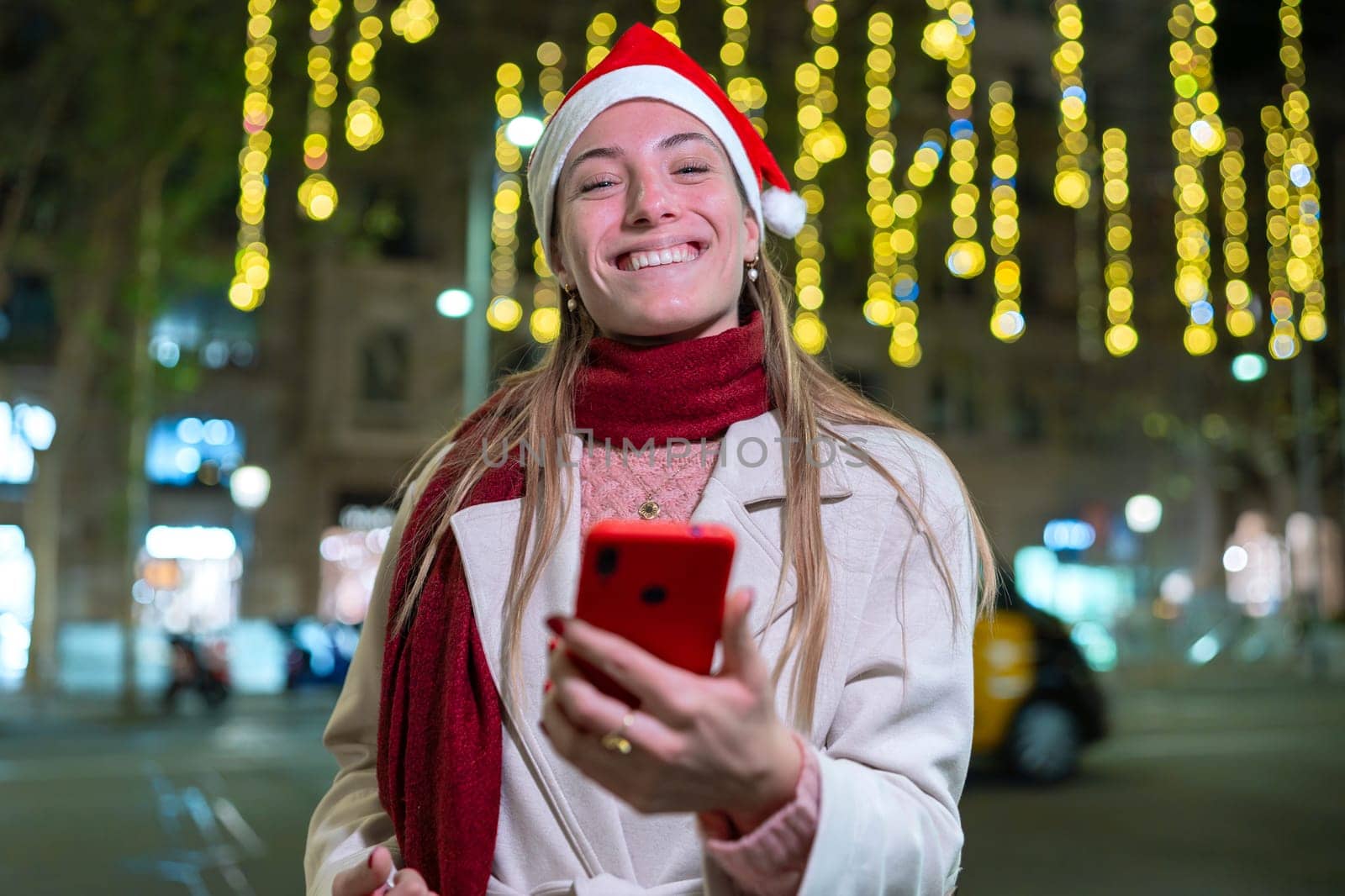 Young woman wearing a Santa hat and red scarf, smiling and holding her smartphone. She stands outdoors at night with festive holiday lights in the background, creating a joyful atmosphere.