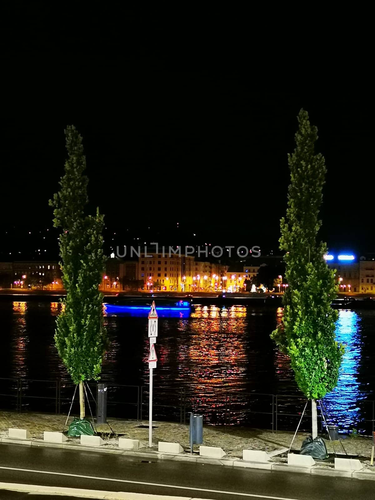The night showcases vibrant reflections on the river as colorful lights illuminate the buildings, and a boat sails past, creating a picturesque waterfront setting.