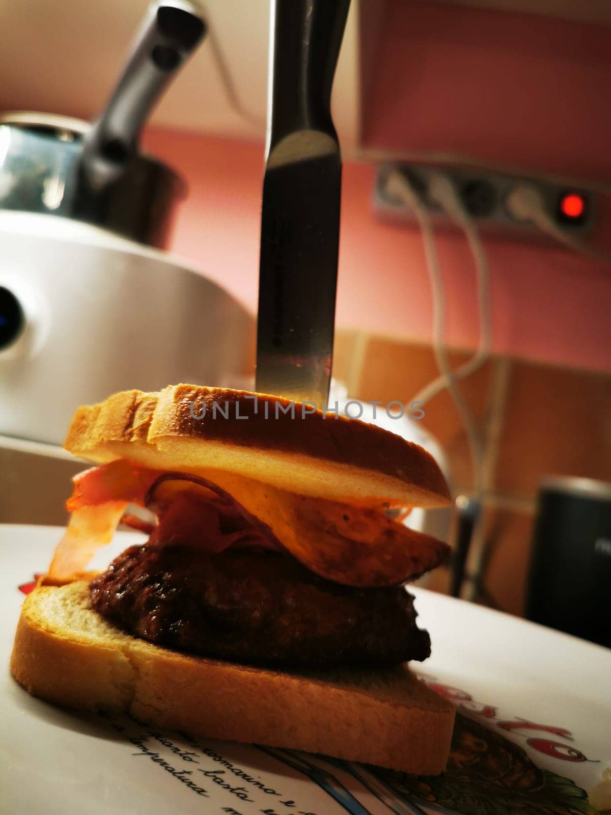 A juicy beef patty topped with crispy bacon is sandwiched between slices of bread, with a knife lodged upright, all set against a home kitchen background.
