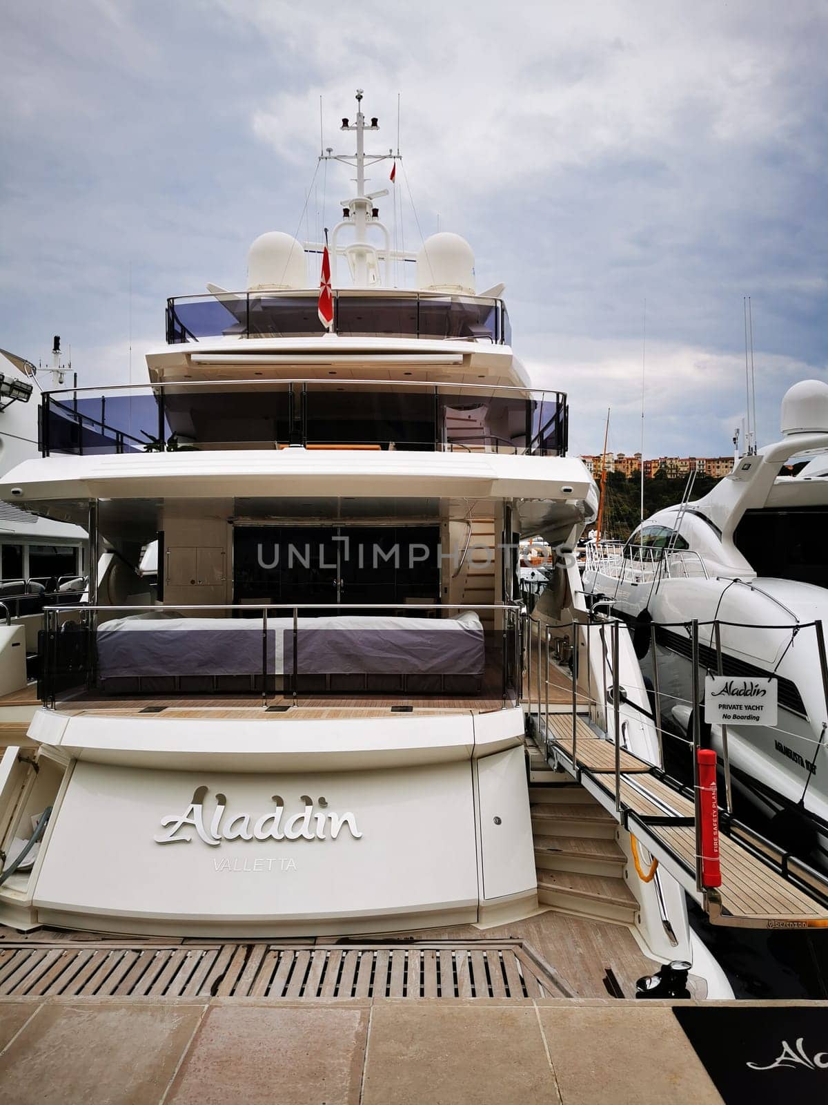 The luxury yacht Aladdin is moored at a marina with fellow vessels nearby, showcasing its elegant design under a cloudy afternoon sky in a coastal city.