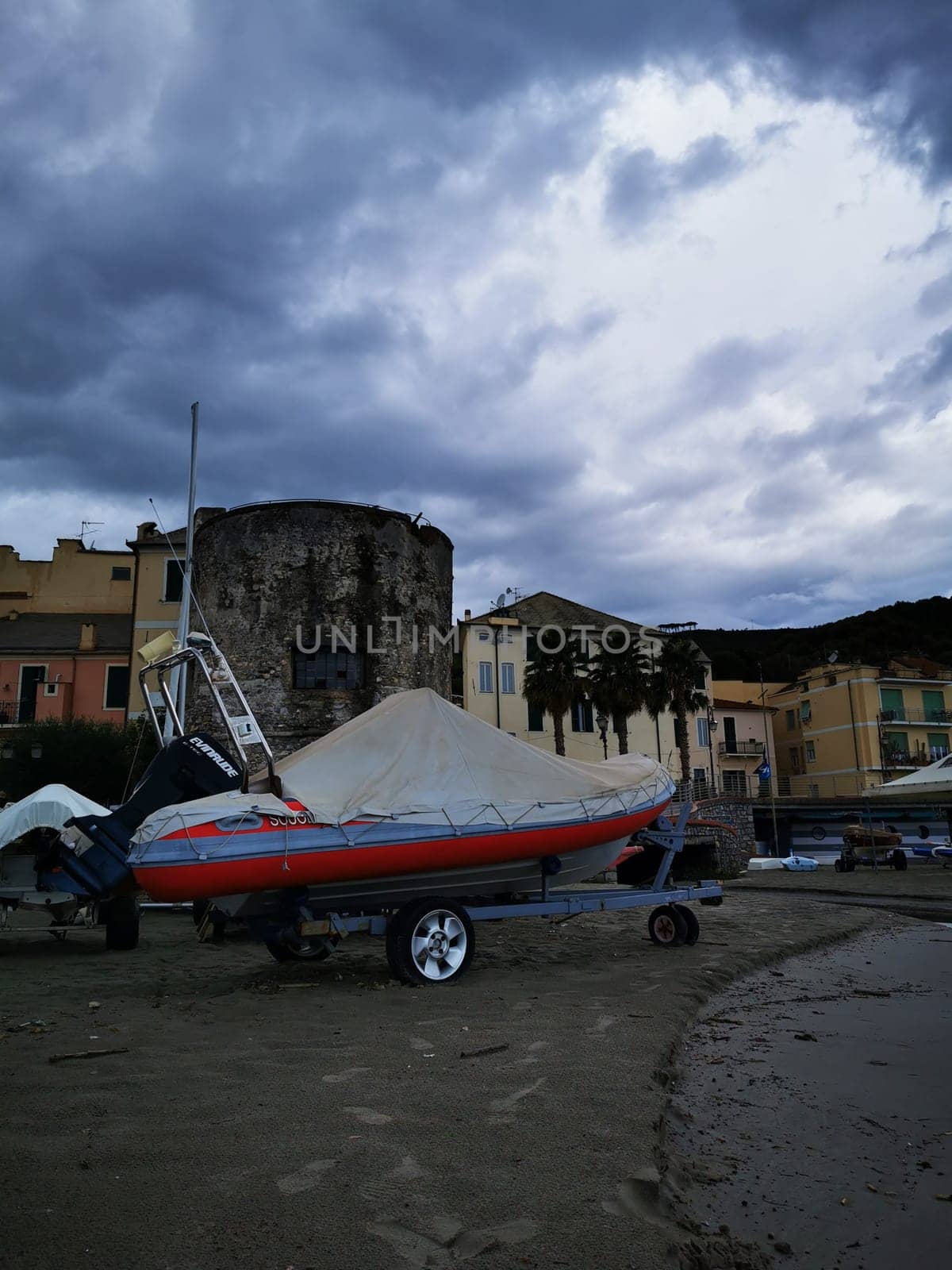 A colorful boat is parked on the sandy beach near a historical stone tower, while dark clouds linger over the seaside village, creating a moody atmosphere.