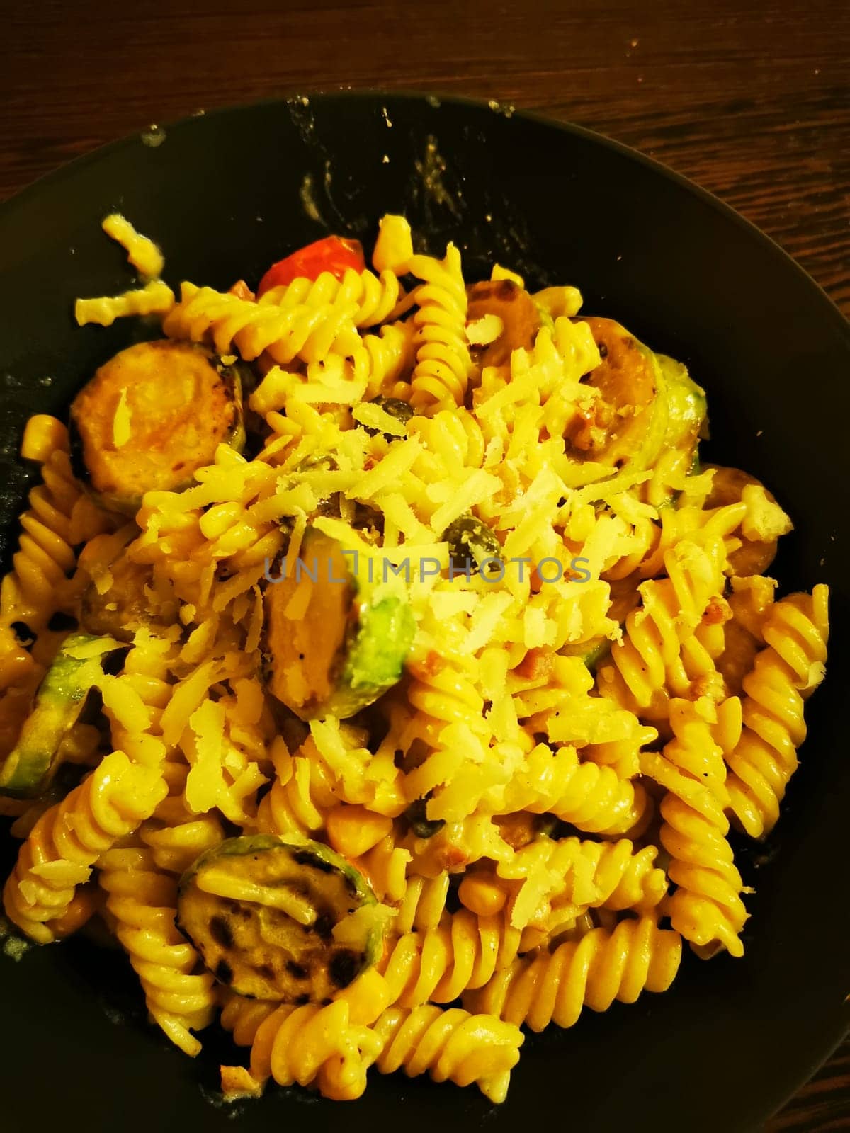 A vibrant dish featuring spiral pasta tossed with zucchini, bell peppers, and a sprinkle of cheese, served in a black bowl against a rustic wooden backdrop.