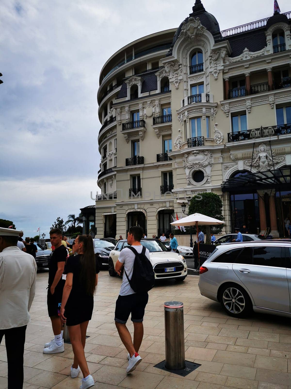 Groups of individuals stroll elegantly along a vibrant street near a grand architectural structure, surrounded by stylish cars and a lively atmosphere.
