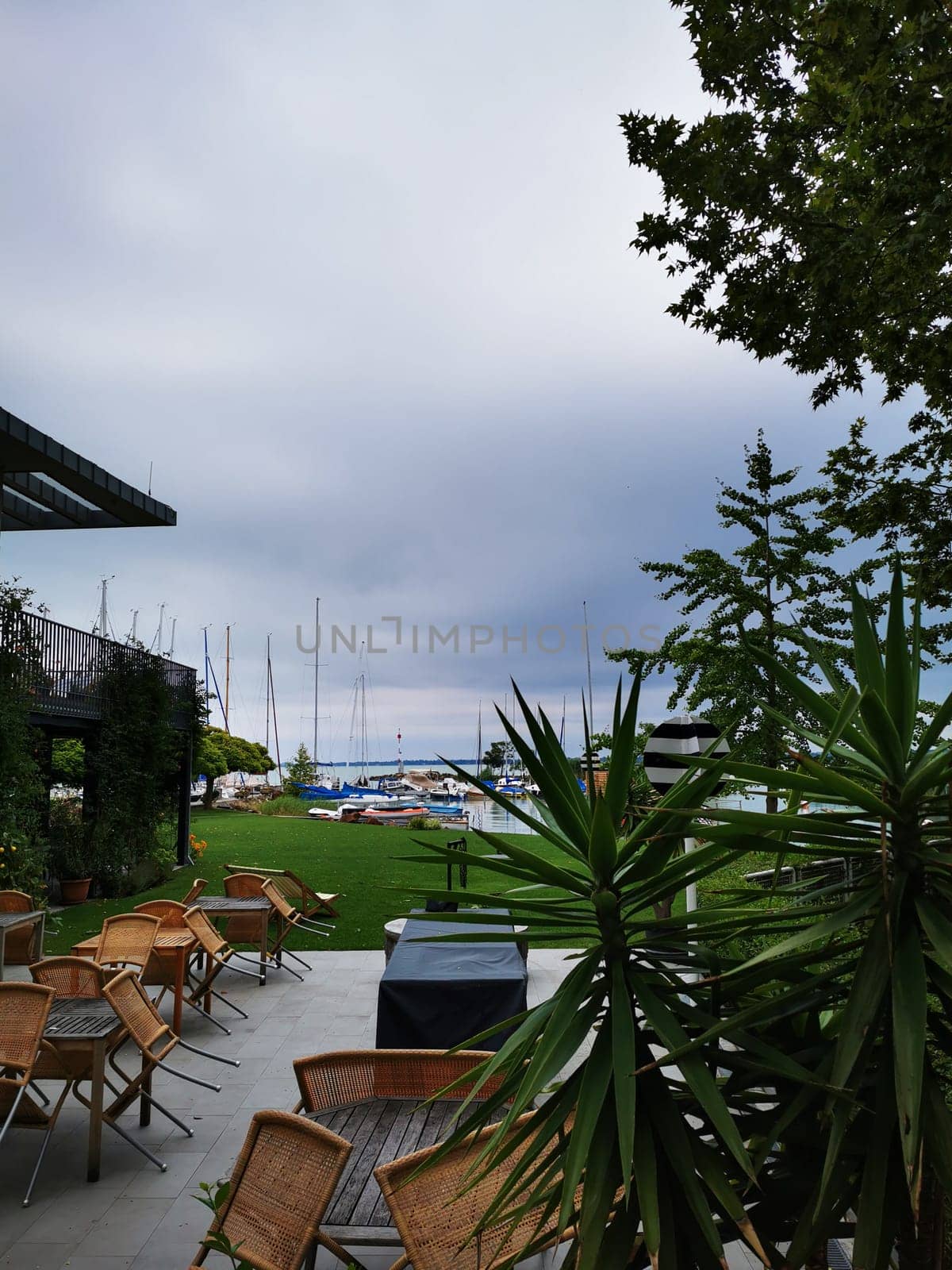 Visitors relax in a tranquil outdoor seating area overlooking a marina filled with sailboats, surrounded by lush greenery under a cloudy sky.