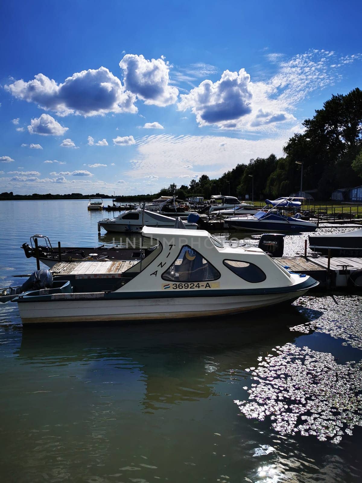 Several boats are moored at a tranquil marina, reflecting on the water's surface. The sky is bright and dotted with fluffy clouds, creating a picturesque setting.