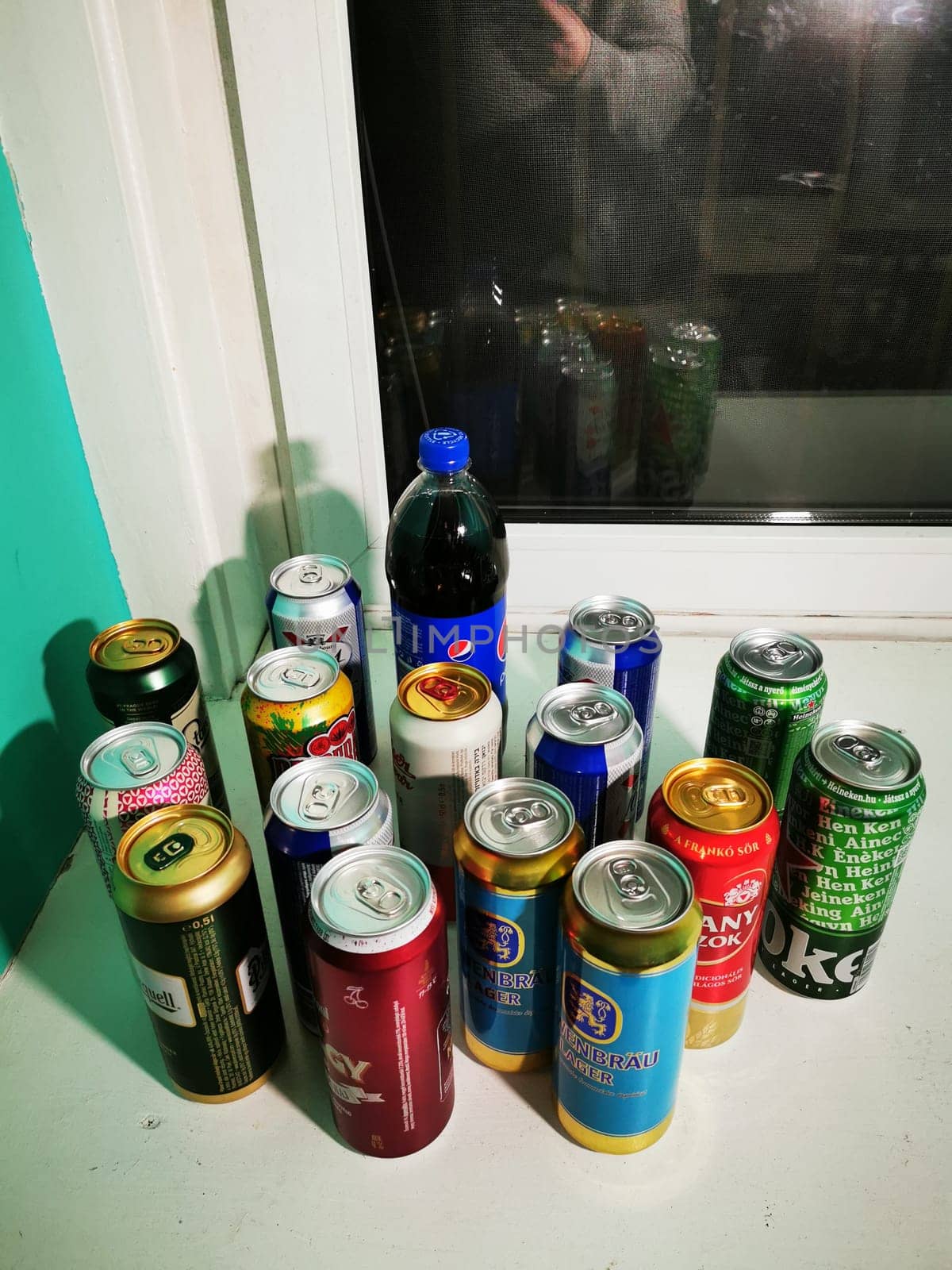A variety of beverage cans and a bottle positioned on a windowsill, showcasing different colors and labels in a cozy, brightly painted room.