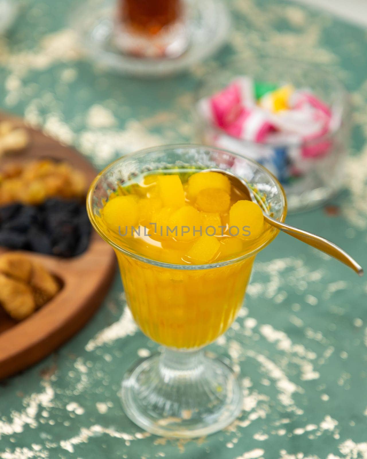 Glass of quince dessert is sitting on a table with a spoon in it, next to a plate of dried fruits and a cup of turkish tea