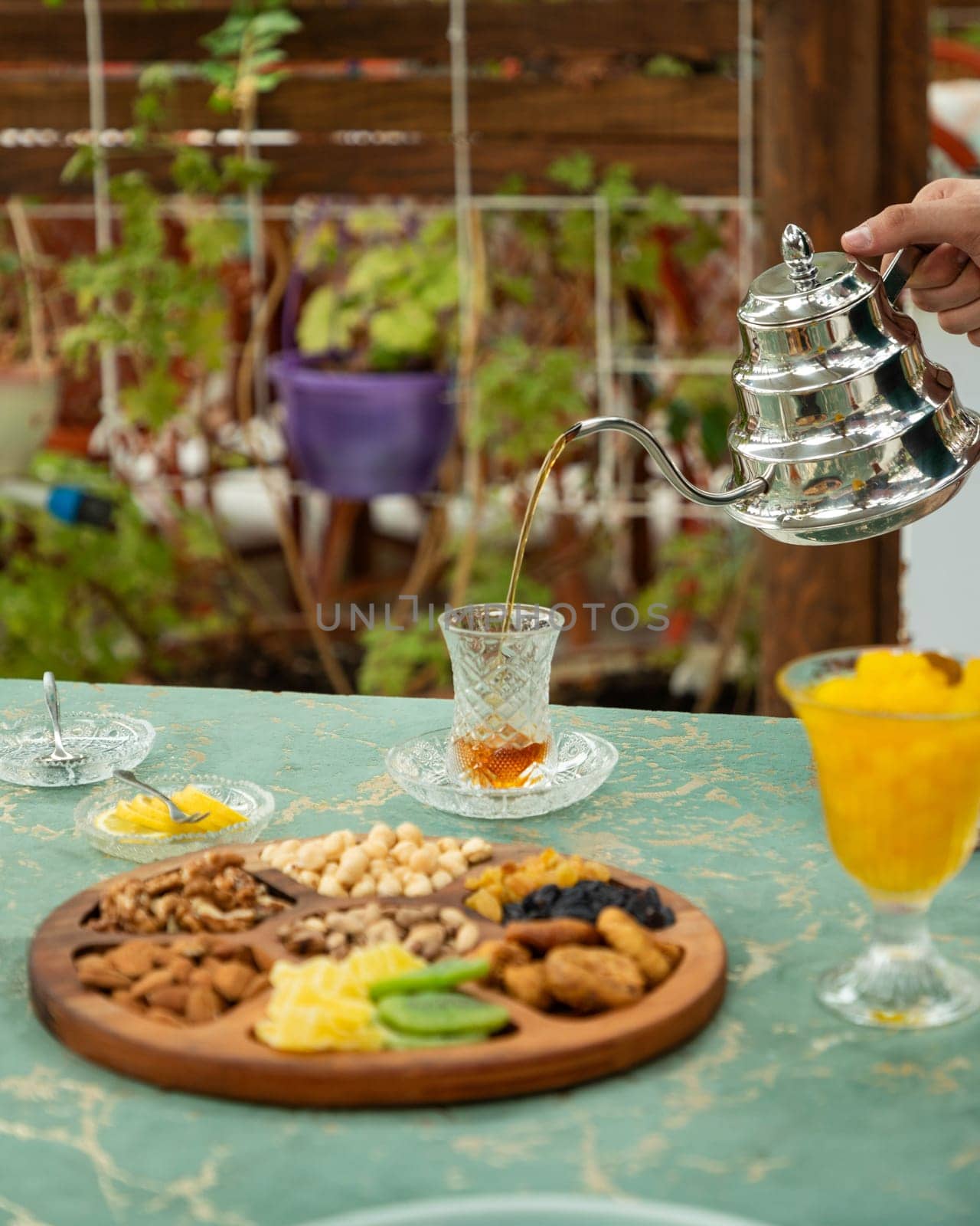 Person pouring tea from a traditional turkish teapot into a glass on a table with a wooden tray of nuts and dried fruits