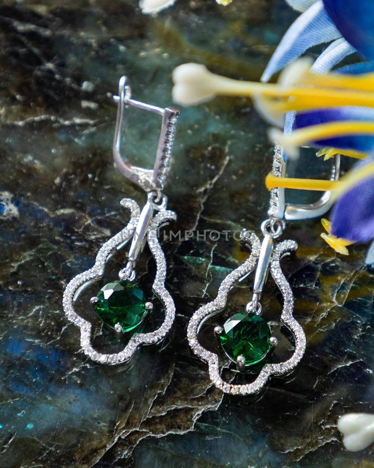 Pair of emerald and diamond earrings are lying on a stone surface with a blue flower next to them
