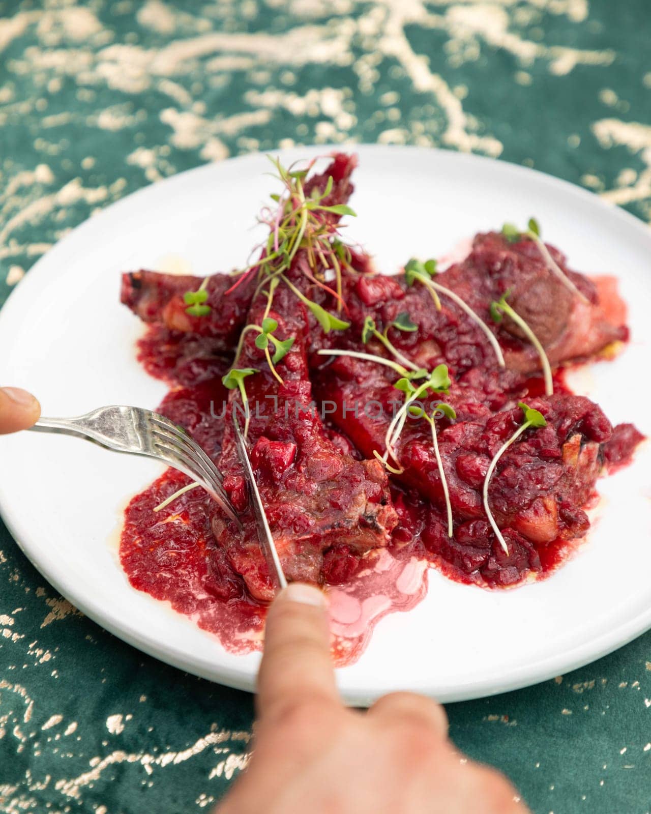 Grilled lamb ribs with cranberry sauce and microgreens are being cut by a chef on a white plate